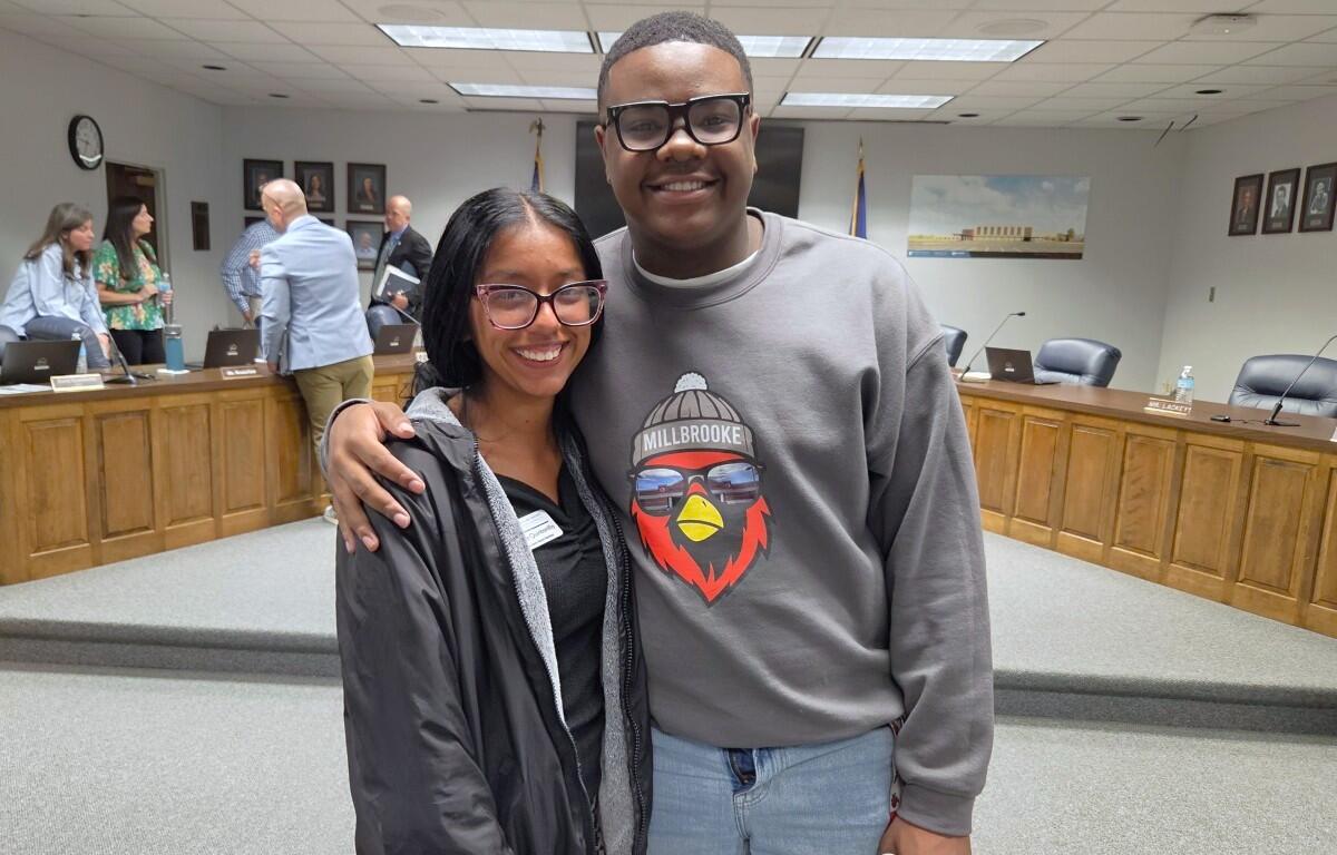 Two friends pose together in a formal meeting room, smiling at the camera with wood desks and people in the background.