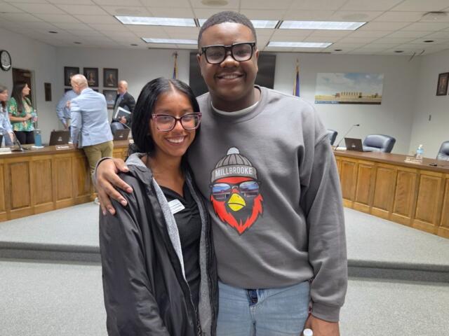 Two friends pose together in a formal meeting room, smiling at the camera with wood desks and people in the background.