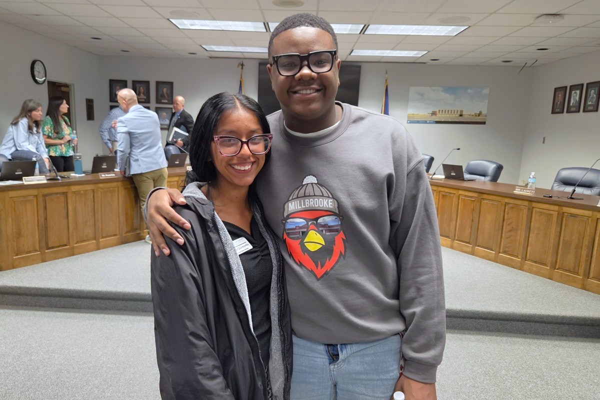 Two friends pose together in a formal meeting room, smiling at the camera with wood desks and people in the background.