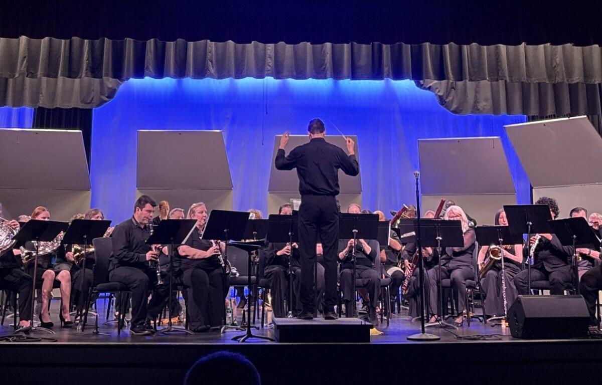 Conductor in black leads a large orchestra on a stage with blue backdrop and sheet music stands.