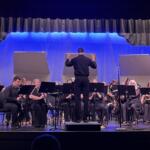 Conductor in black leads a large orchestra on a stage with blue backdrop and sheet music stands.