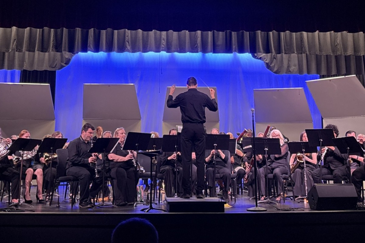 Conductor in black leads a large orchestra on a stage with blue backdrop and sheet music stands.