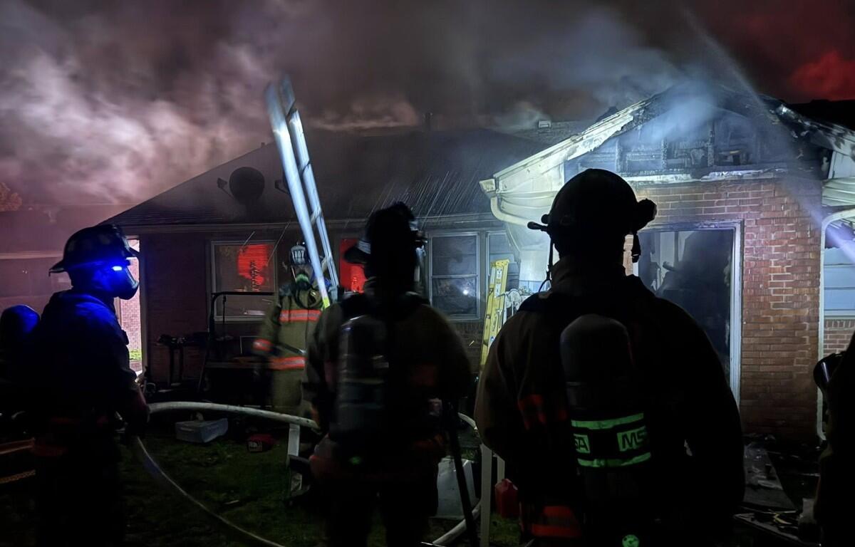 Firefighters in turnout gear battle a house fire at night, smoke billowing from the roof with a ladder leaning against the building.