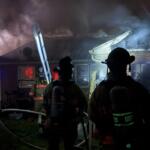 Firefighters in turnout gear battle a house fire at night, smoke billowing from the roof with a ladder leaning against the building.