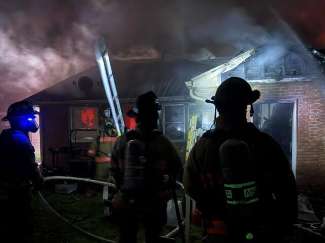 Firefighters in turnout gear battle a house fire at night, smoke billowing from the roof with a ladder leaning against the building.