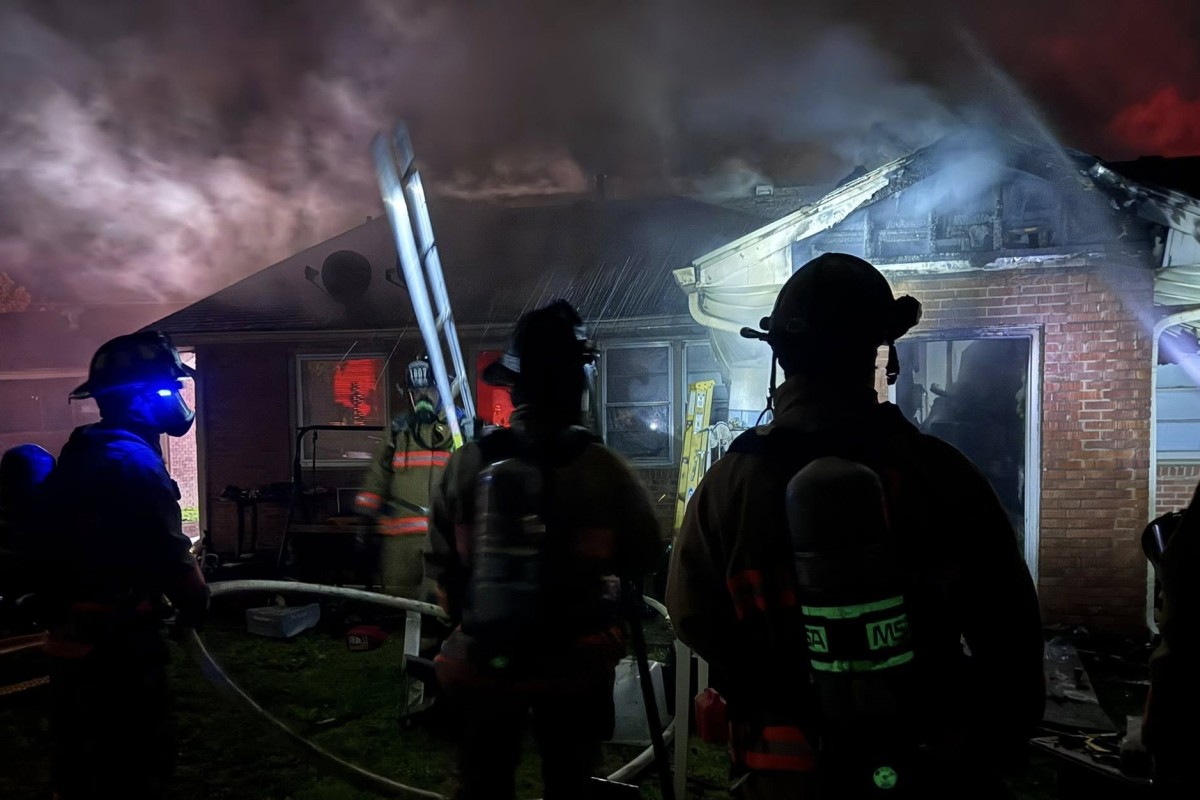 Firefighters in turnout gear battle a house fire at night, smoke billowing from the roof with a ladder leaning against the building.