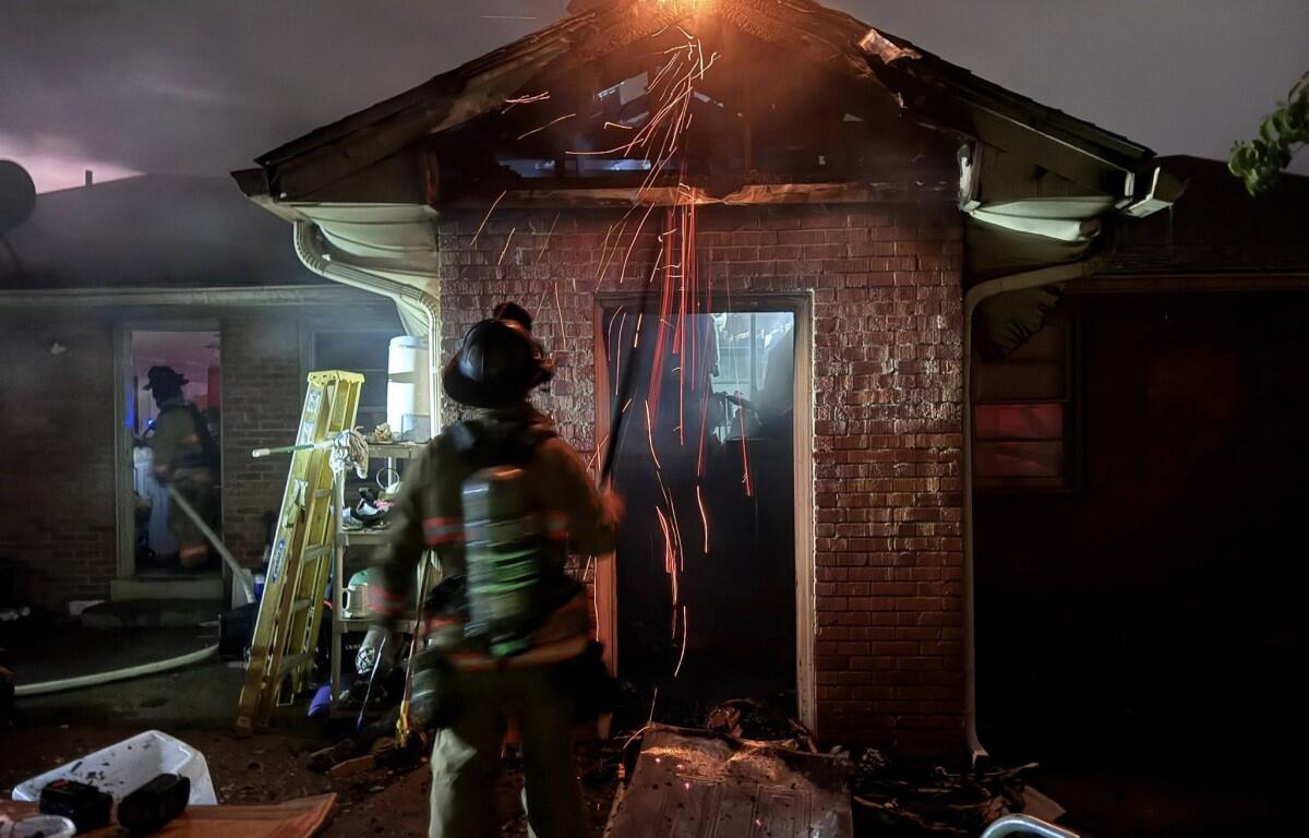 Firefighter in full turnout gear at night in front of a burning brick house; sparks fly from the damaged roof area.
