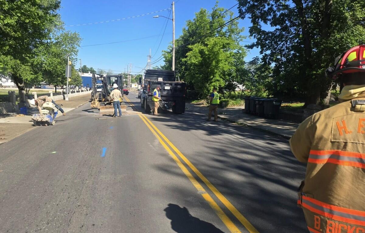 Construction crew working on a sunlit street with a backhoe, dump trucks, and workers in high-visibility vests.