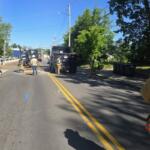 Construction crew working on a sunlit street with a backhoe, dump trucks, and workers in high-visibility vests.