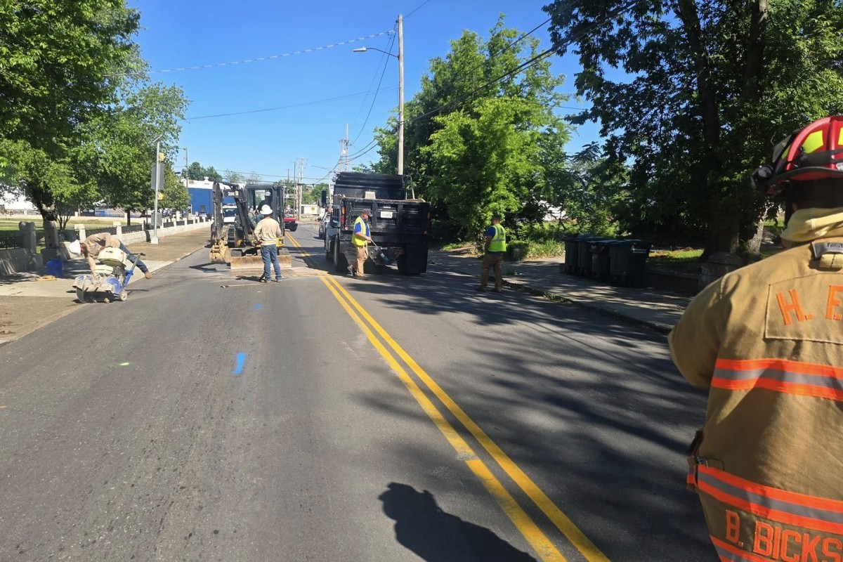 Construction crew working on a sunlit street with a backhoe, dump trucks, and workers in high-visibility vests.