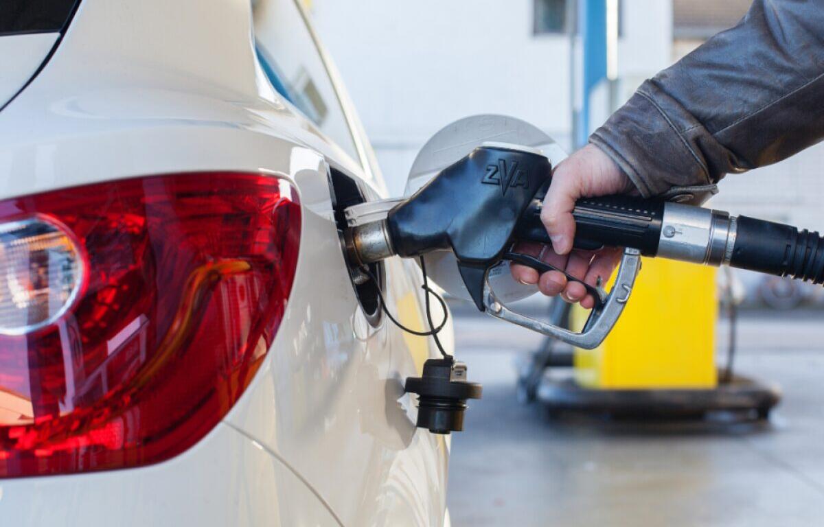 Person refueling a white car at a gas station, inserting a fuel nozzle into the rear fuel filler area.