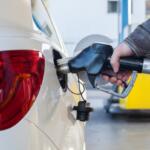 Person refueling a white car at a gas station, inserting a fuel nozzle into the rear fuel filler area.