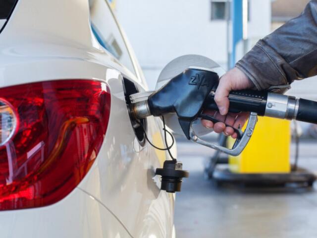 Person refueling a white car at a gas station, inserting a fuel nozzle into the rear fuel filler area.