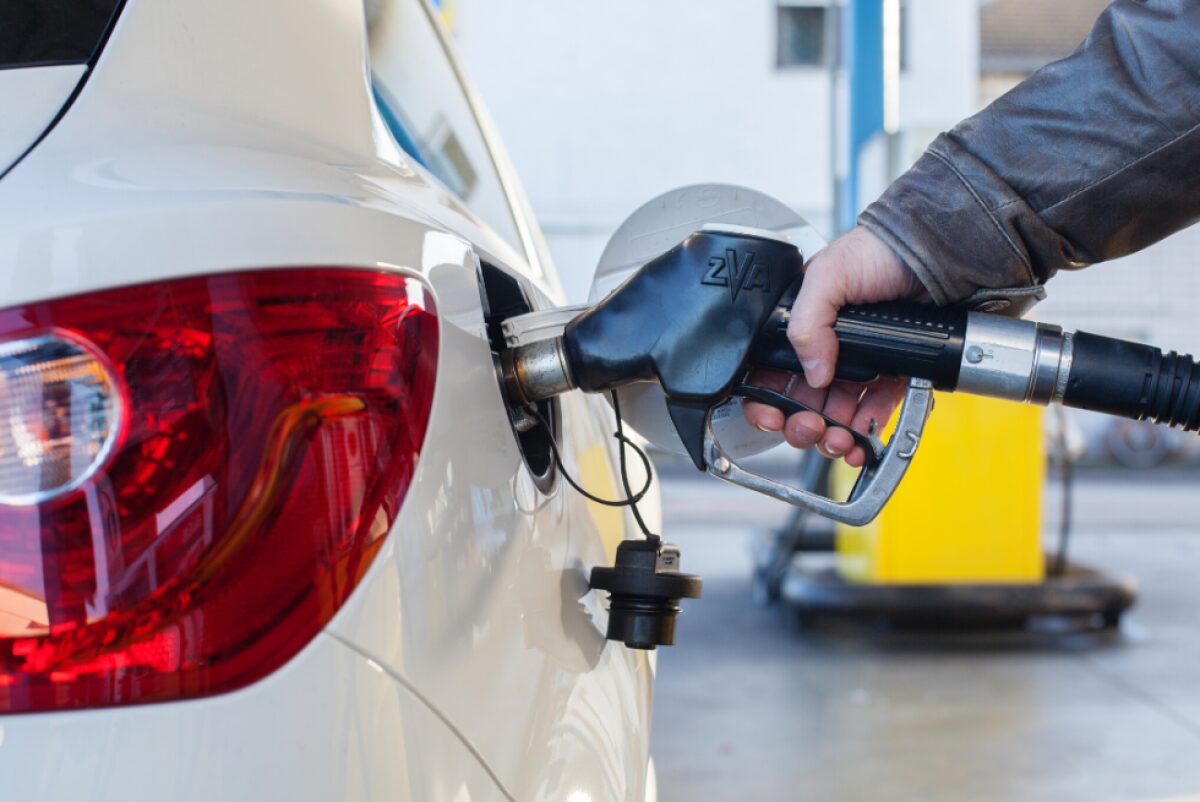 Person refueling a white car at a gas station, inserting a fuel nozzle into the rear fuel filler area.