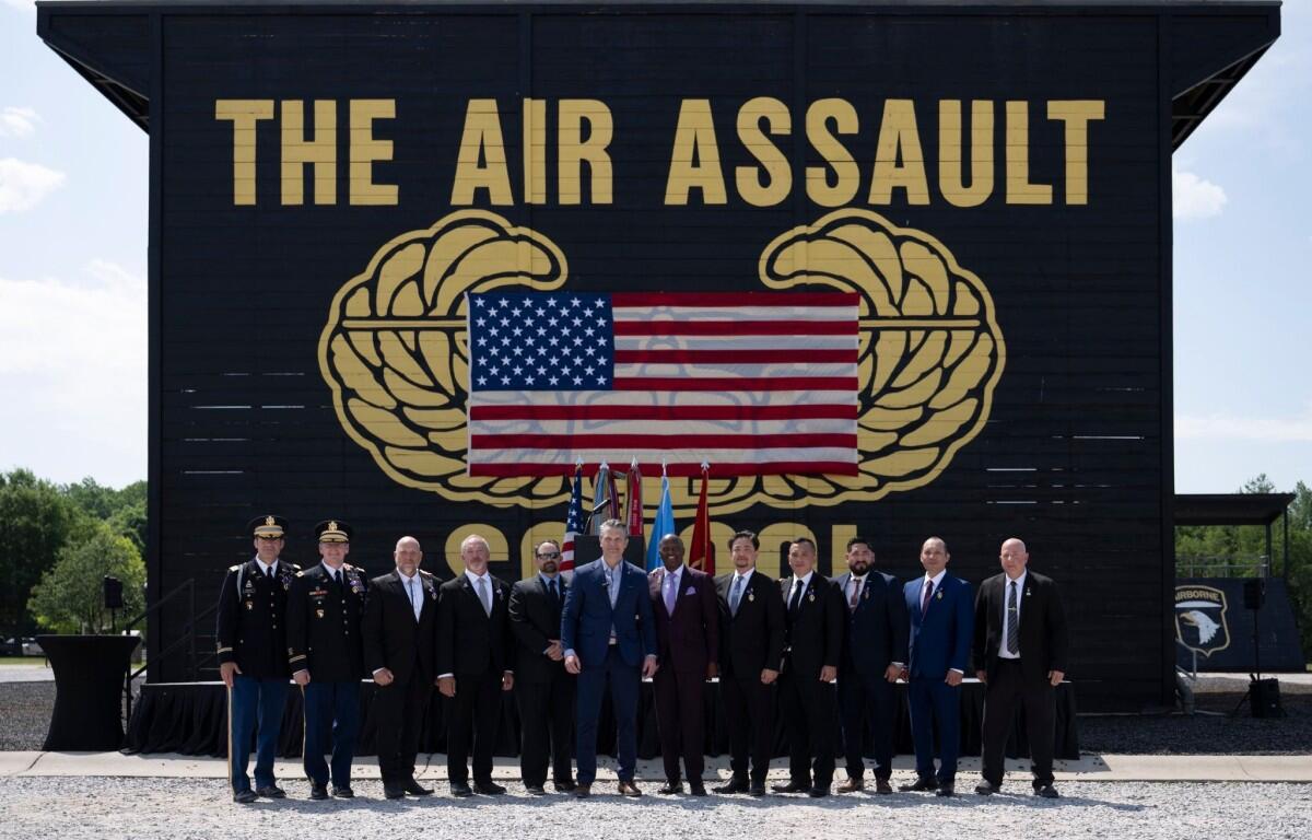 Group of military personnel and civilians in formal attire posing outdoors in front of a large black backdrop that reads THE AIR ASSAULT with a U.S. flag displayed between gold insignia.
