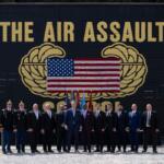 Group of military personnel and civilians in formal attire posing outdoors in front of a large black backdrop that reads THE AIR ASSAULT with a U.S. flag displayed between gold insignia.