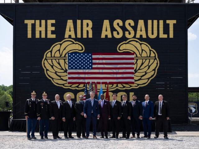 Group of military personnel and civilians in formal attire posing outdoors in front of a large black backdrop that reads THE AIR ASSAULT with a U.S. flag displayed between gold insignia.