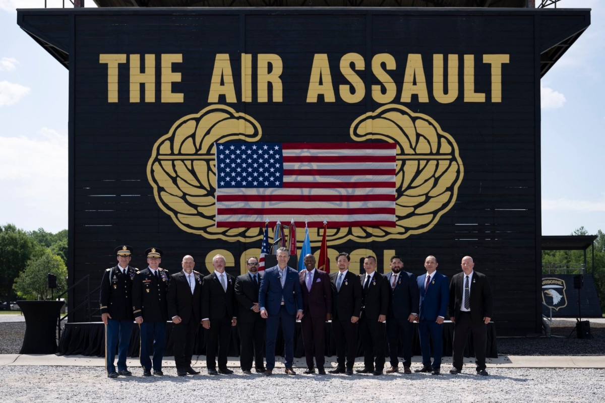 Group of military personnel and civilians in formal attire posing outdoors in front of a large black backdrop that reads THE AIR ASSAULT with a U.S. flag displayed between gold insignia.