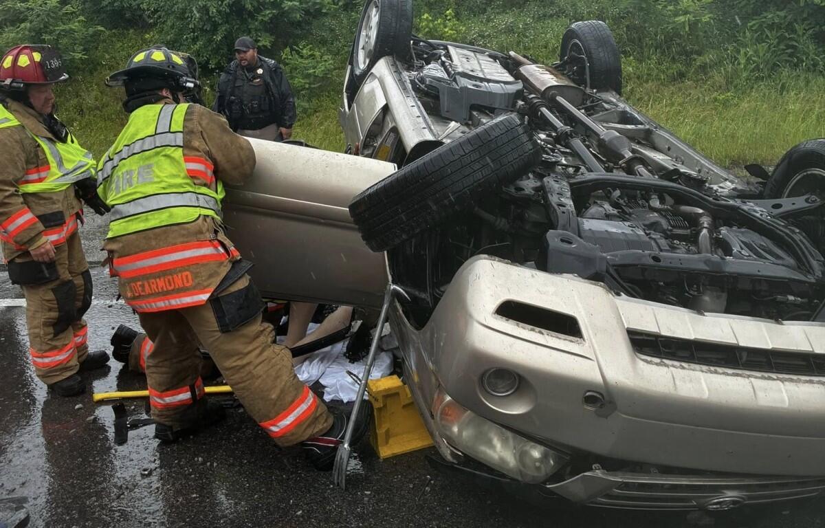 Firefighters in turnout gear stabilize a rolled SUV on a wet road during a vehicle rescue.