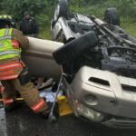 Firefighters in turnout gear stabilize a rolled SUV on a wet road during a vehicle rescue.