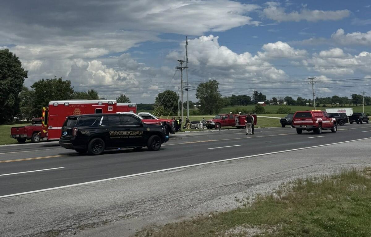 Emergency responders and multiple emergency vehicles on a rural highway shoulder around a crash, including a sheriff SUV, a red ambulance, and fire trucks, with bystanders nearby.