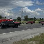 Emergency responders and multiple emergency vehicles on a rural highway shoulder around a crash, including a sheriff SUV, a red ambulance, and fire trucks, with bystanders nearby.