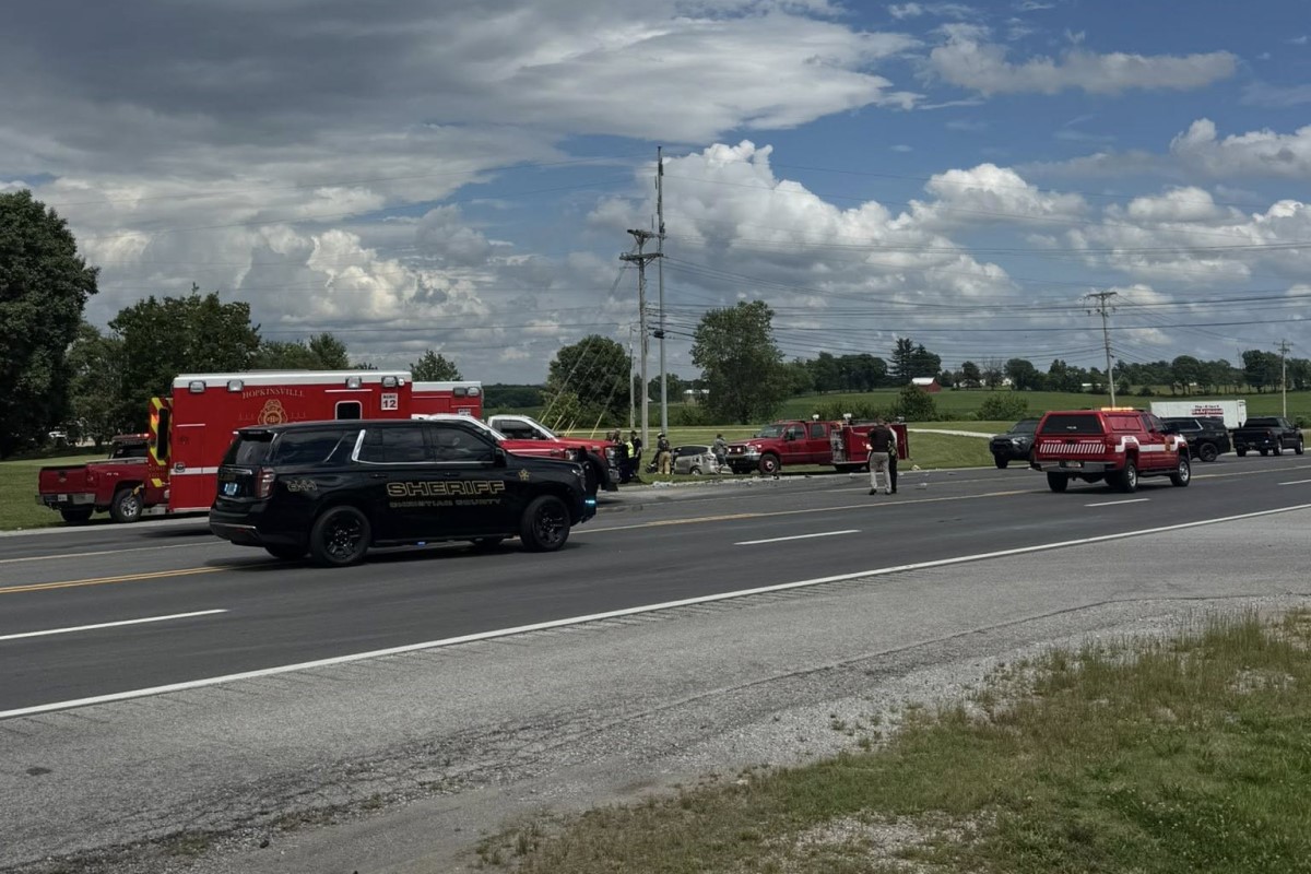 Emergency responders and multiple emergency vehicles on a rural highway shoulder around a crash, including a sheriff SUV, a red ambulance, and fire trucks, with bystanders nearby.
