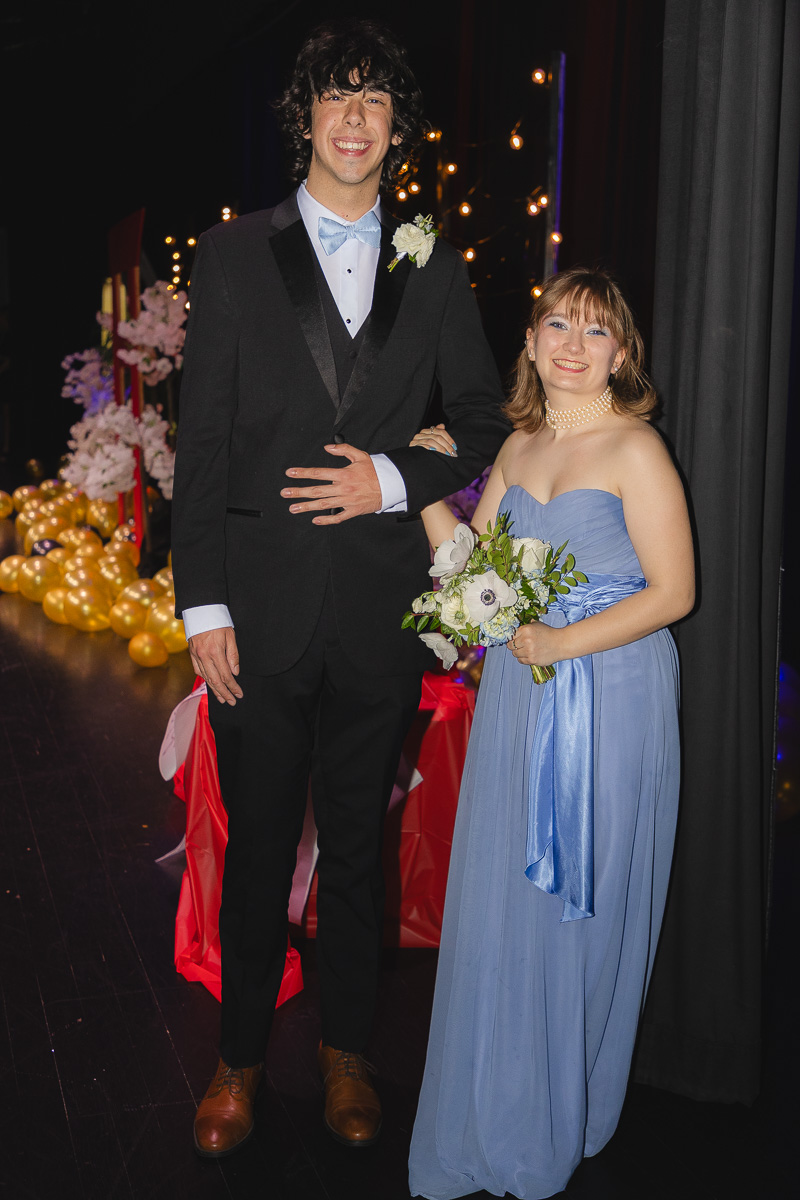 Smiling couple in formal wear posing onstage: man in a black tux with a light blue bow tie and boutonniere, woman in a blue strapless gown holding a white bouquet.