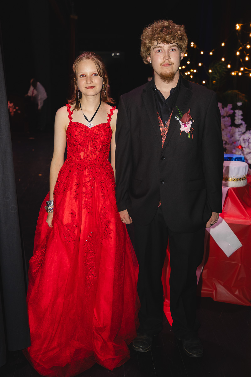 Couple at a formal event: woman in a red lace ball gown and man in a black suit with a boutonniere, standing together outdoors at night with fairy lights.