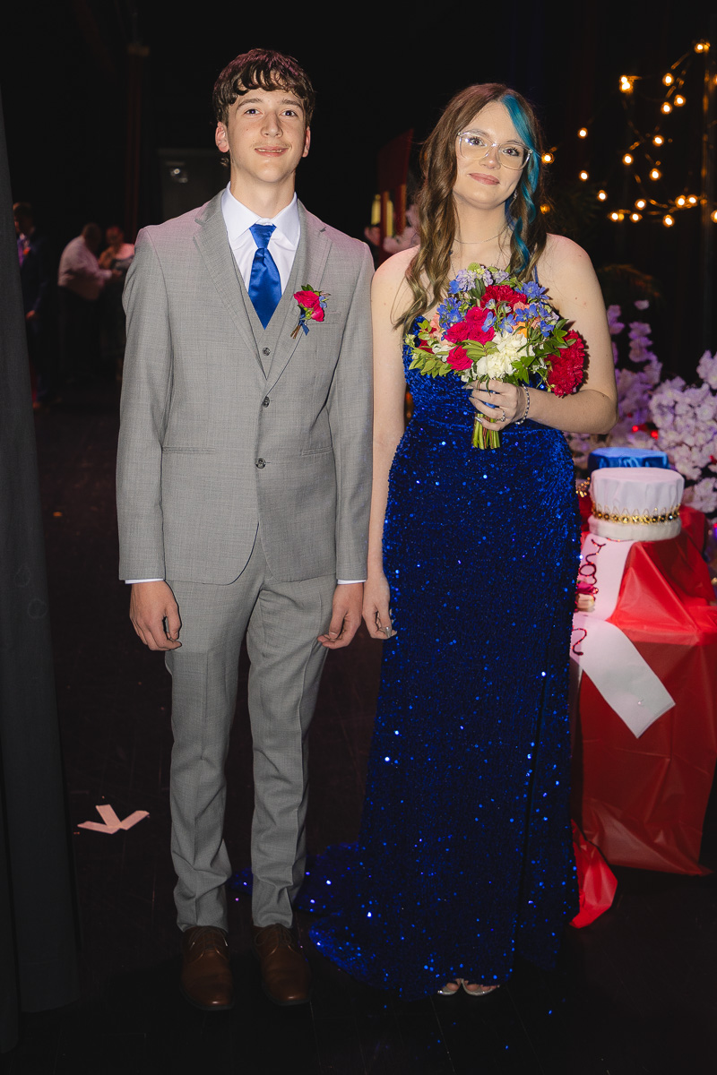 Two teens in formal attire pose together at a party: a guy in a gray suit and blue tie, and a girl in a blue sequined gown holding a bouquet.