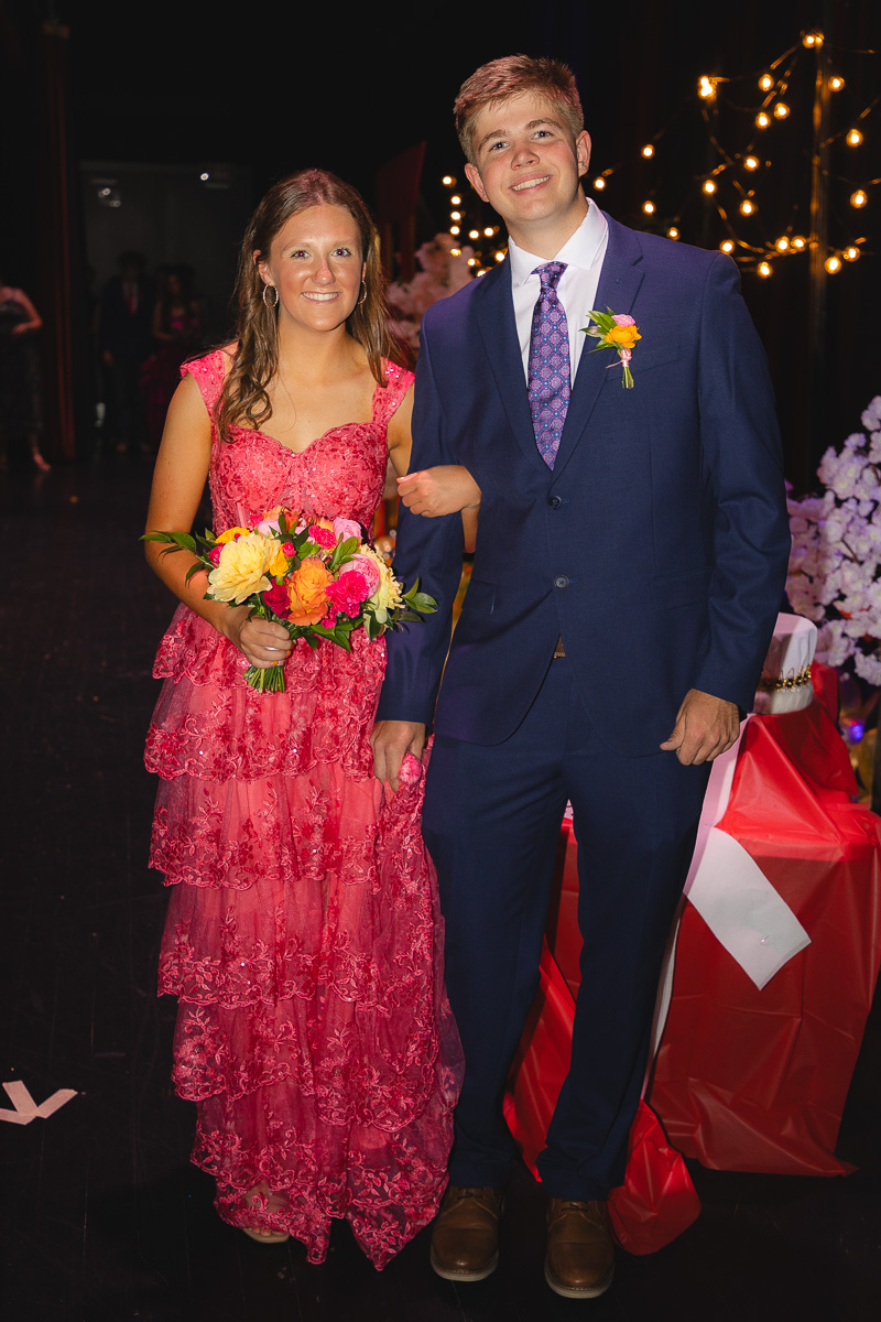 Young couple at a formal event walking arm-in-arm; the woman wears a pink lace gown and holds a colorful bouquet, the man in a navy suit with a purple tie and boutonniere smiles at the camera.