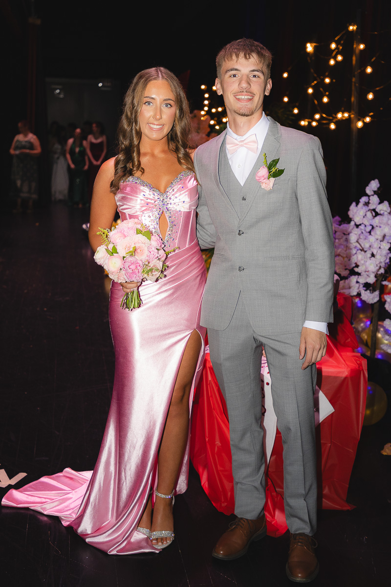 Couple in formal wear posing at a prom, girl in pink satin gown holding a bouquet and the boy in a gray suit with a pink boutonniere.