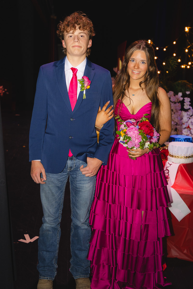 A young couple posing at a formal event: he in a blue blazer and pink tie, she in a magenta tiered dress holding a colorful bouquet.