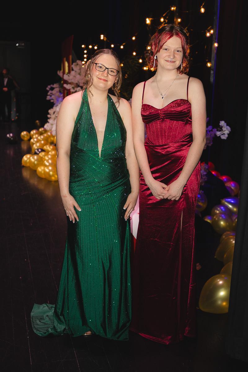 Two young women in formal gowns pose for a photo at a festive event; one in a green sequin dress, the other in a burgundy velvet gown, with string lights and balloons in the background.