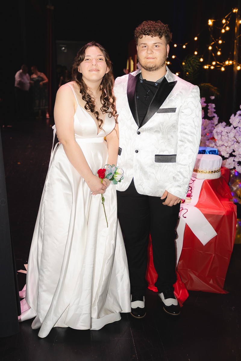 Couple in formal attire posing at a party, the woman in a white gown holding a rose bouquet and the man in a patterned white jacket.