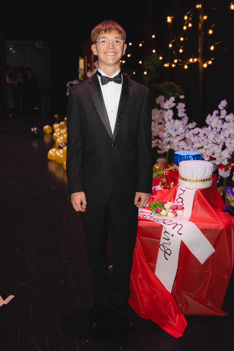 Young man in a black tuxedo and bow tie posing at a festive event with decorative lights and a wrapped cake on a red table nearby