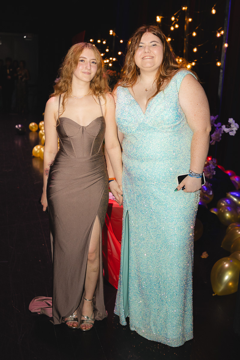 Two women in formal gowns smile at a party; one in a taupe gown with a thigh slit, the other in a light blue sequin dress, holding hands amid string lights and balloons.