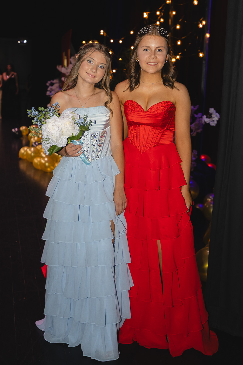 Two teenage girls in formal gowns pose together at a party, one in a light blue tiered dress holding a bouquet, the other in a red tiered gown with a slit.