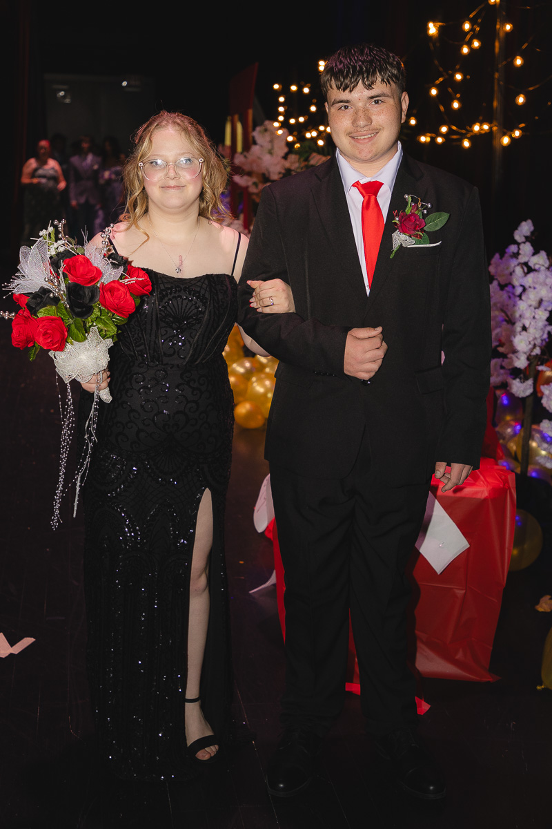 Young couple in formal wear walk arm in arm at a prom-like event; she holds a red rose bouquet and wears a black sequined gown with a slit, he wears a black suit with a red tie and boutonniere.