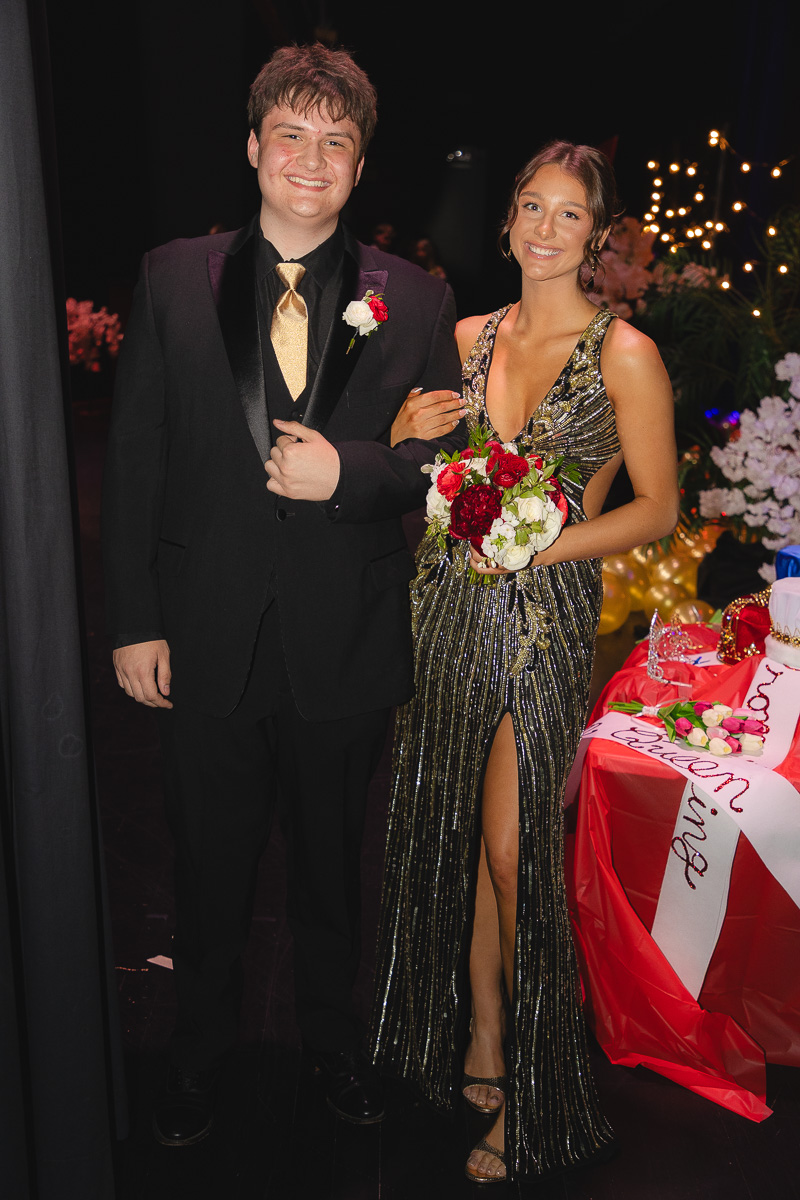 Young man in a black suit and gold tie with boutonniere stands next to a smiling young woman in a glittery gown, holding a bouquet at a formal event.