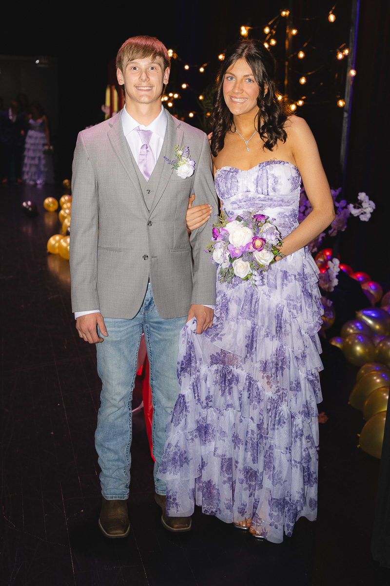 Young couple at a formal event posing together; the man in a gray suit with lavender tie and boutonniere, the woman in a purple bouquet dress holding flowers.