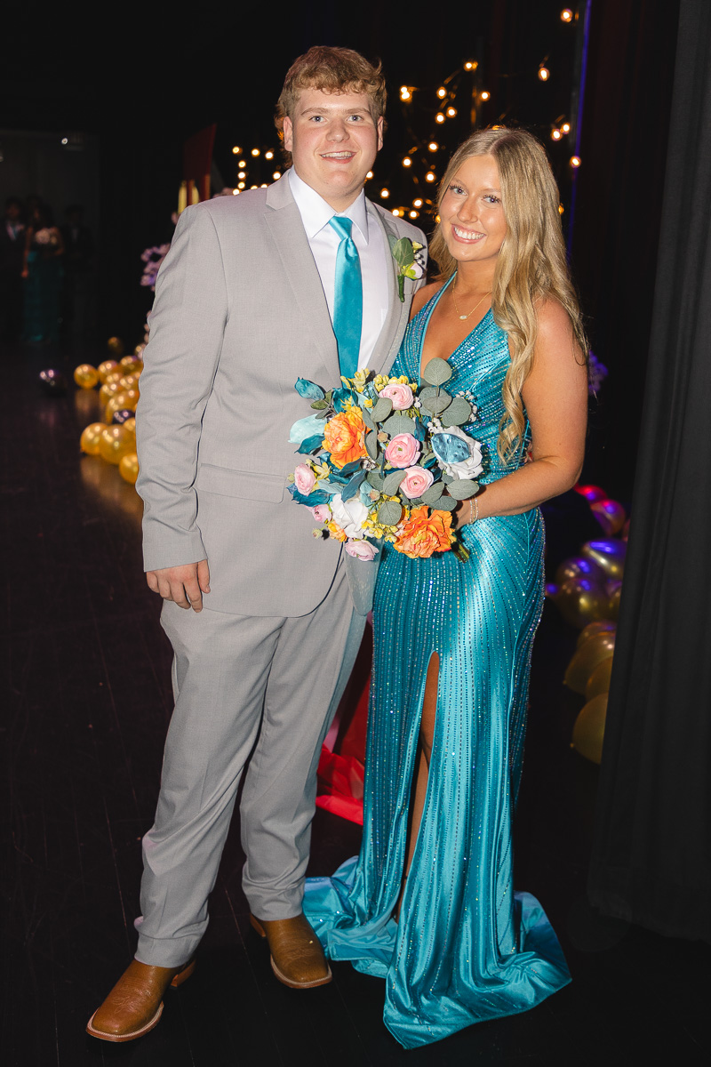 Couple posing at a formal event: man in a light gray suit and teal tie, woman in a turquoise gown holding a colorful bouquet.
