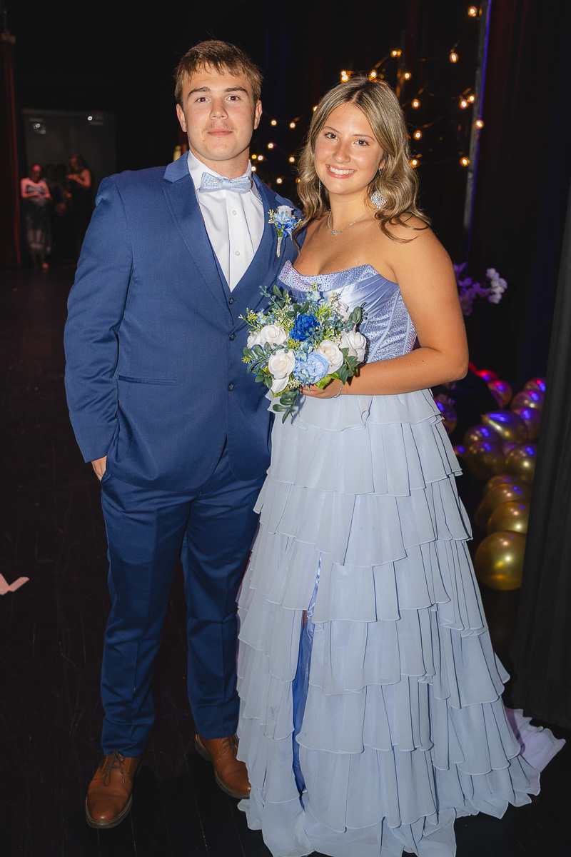 Couple posing at a formal event: man in a blue suit and bow tie, woman in a light blue tiered dress holding a white-blue bouquet.
