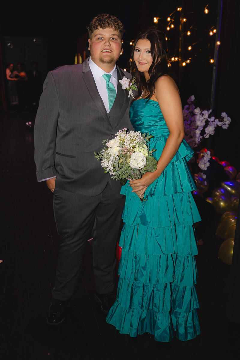 Couple at a formal event posing together; man in a gray suit with teal tie and boutonniere, woman in a turquoise tiered gown holding a white bouquet.