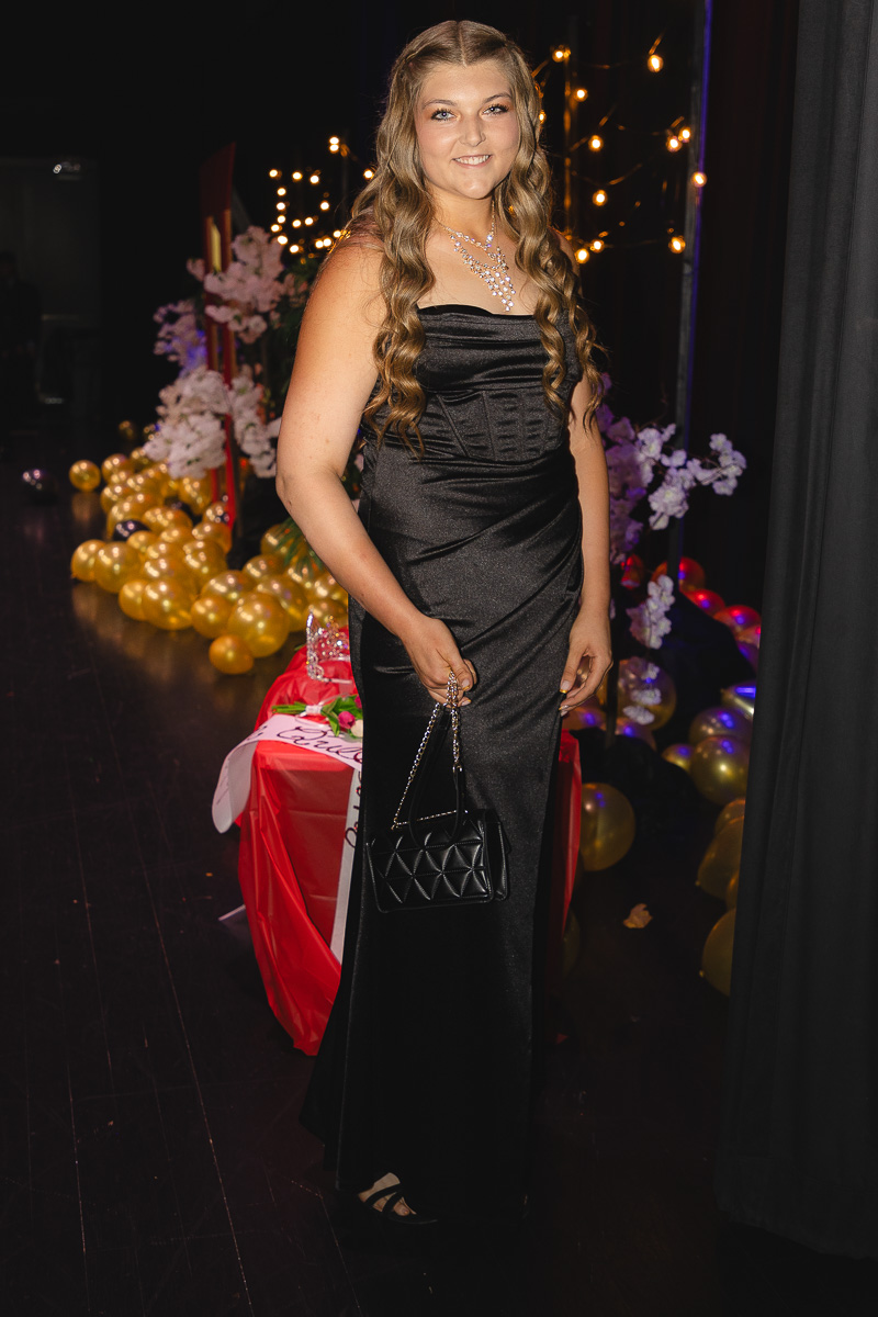 Young woman in a strapless black satin gown with a statement necklace, holding a quilted black purse at a festive event.