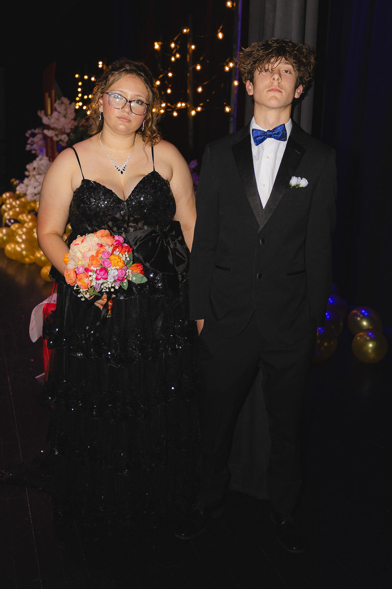 Two teens in formal dress stand together: a girl in a sparkling black gown holding a colorful bouquet, and a boy in a black tuxedo with a blue bow tie.
