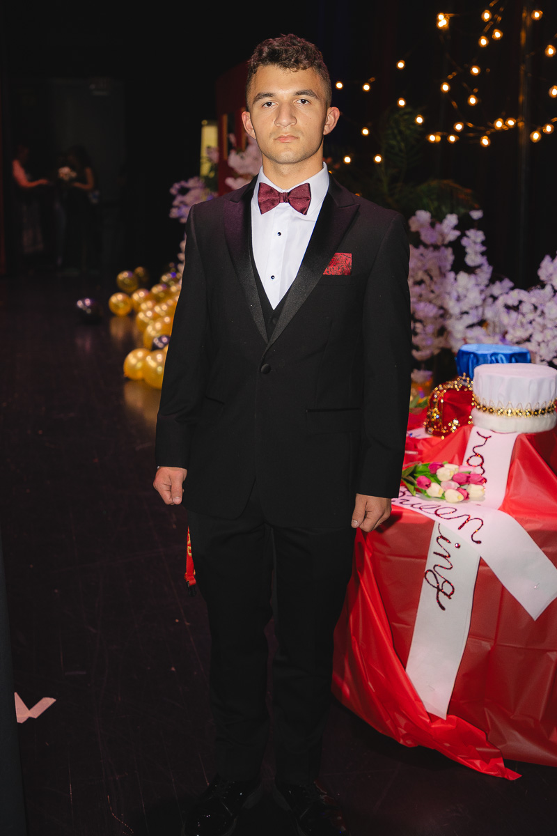 Young man in a black tuxedo with burgundy bow tie posing at a formal event near a decorated dessert table.
