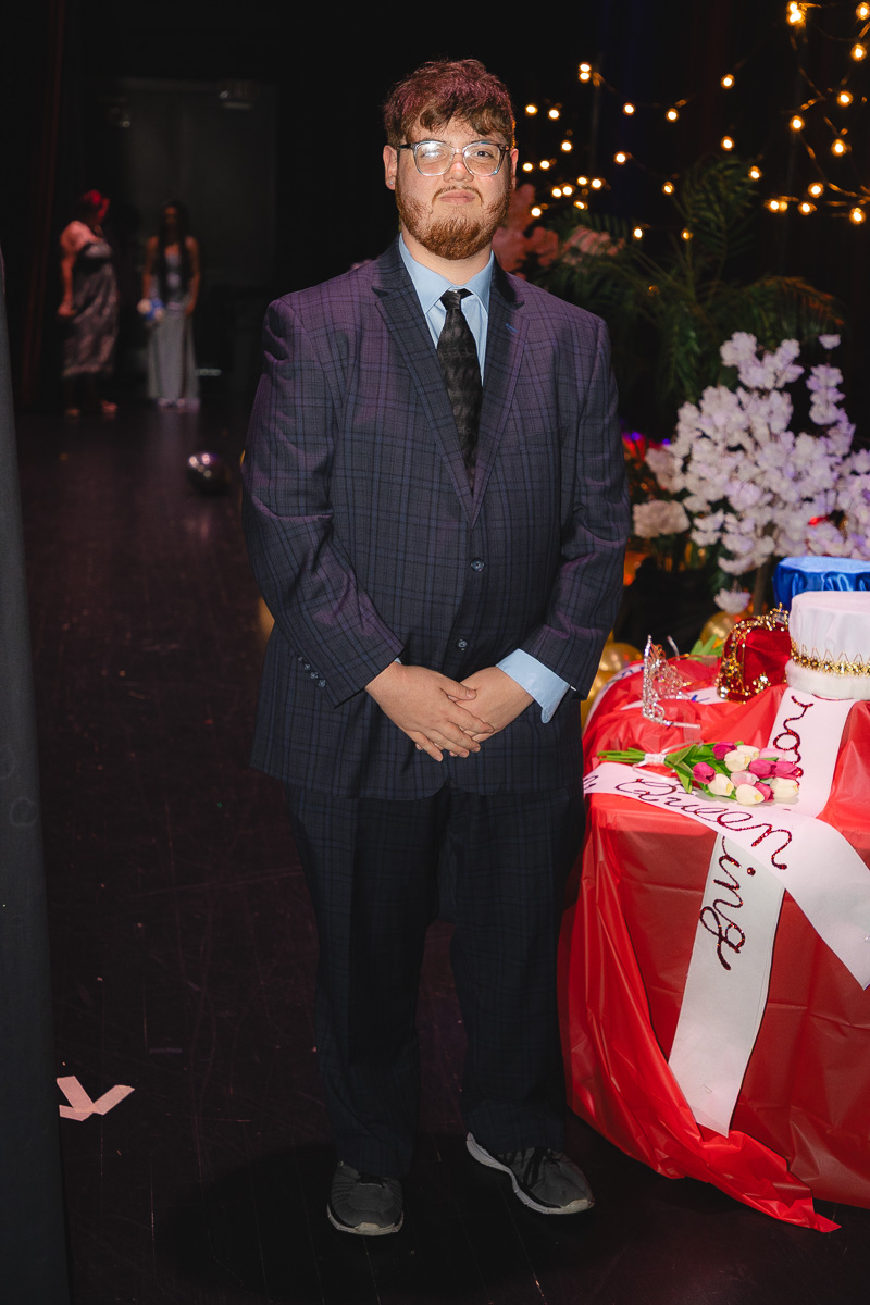 Man in a purple plaid suit and glasses standing beside a decorated table with a cake and flowers at a party.