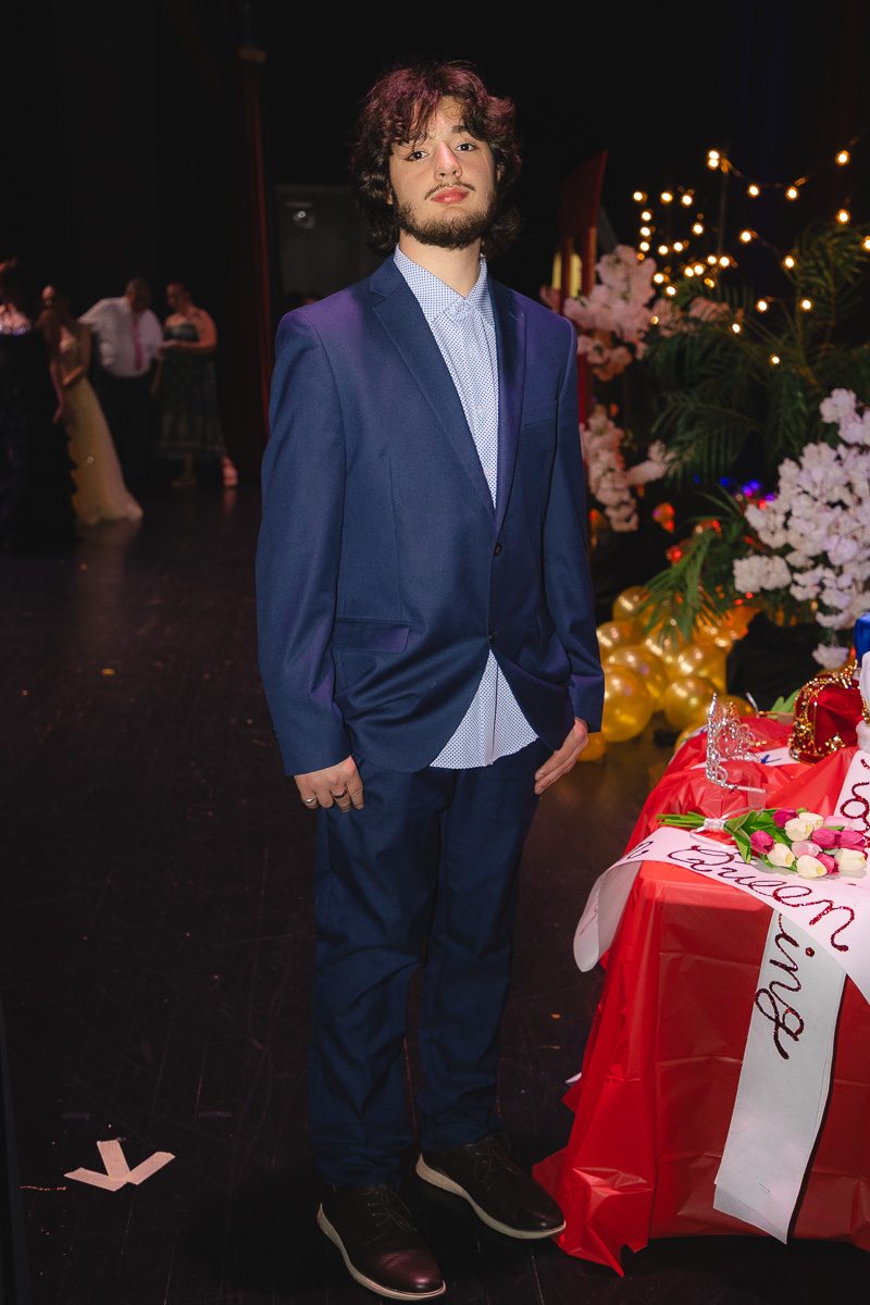 Young man in a navy suit and light blue shirt stands beside a table of gifts and flowers at a formal event.