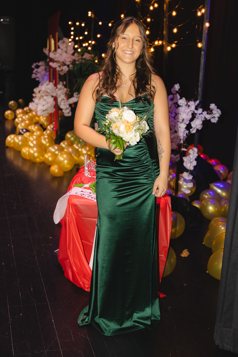 Young woman in a dark green satin gown holding a bouquet of cream and peach flowers, smiling at a festive event with string lights and balloons in the background.